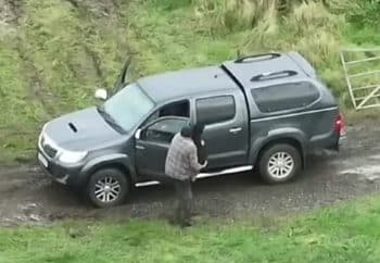 The image shows a gray pickup truck parked on a muddy path in a grassy rural area. A person wearing a jacket and hat is standing by the driver's side door, which is open. There is a metal gate nearby, and the surrounding landscape is lush and green.